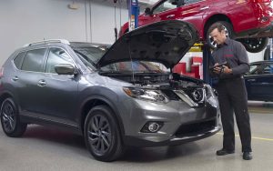 A mechanic working under the open hood of a gray Nissan SUV inside a repair shop.