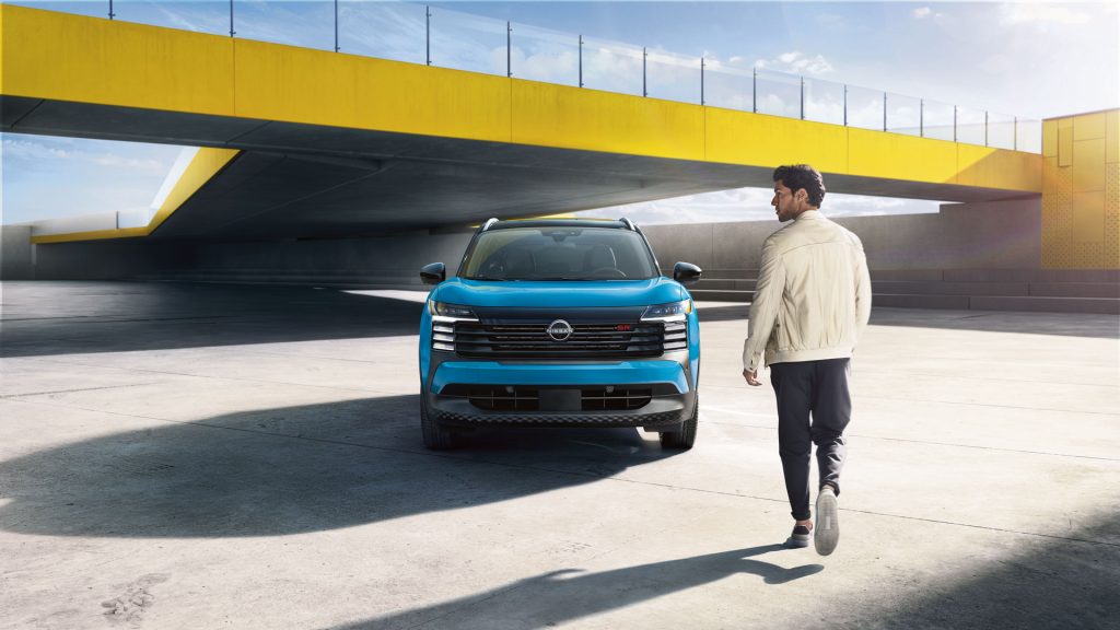 A man strolls away from a blue 2026 Nissan Kicks parked beneath a yellow bridge.