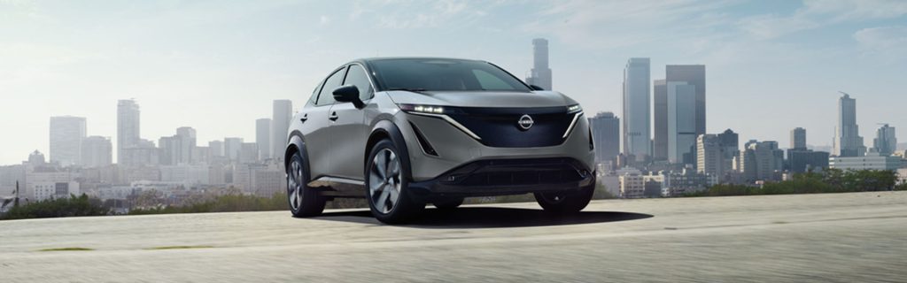 Silver Nissan crossover SUV parked on an elevated surface with a modern city skyline in the background under a clear sky.