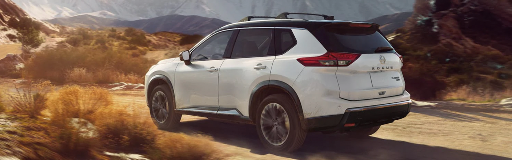 White Nissan Rogue SUV driving on a scenic dirt road with mountains in the background, showcasing its versatility and adventurous design.